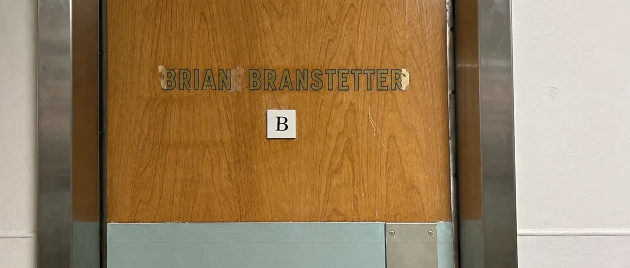 A wood-veneer studio door in a steel frame, the name Brian Branstetter in black stick-on letters and a small B placard, in late warm light